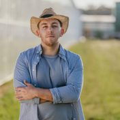 Waist up portrait of a farmer in a hat standing near a greenhouse and looking at the camera